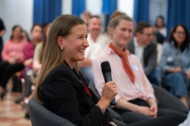 Junge Frau mit Mikrofon in der Hand und Publikum im Hintergrund.